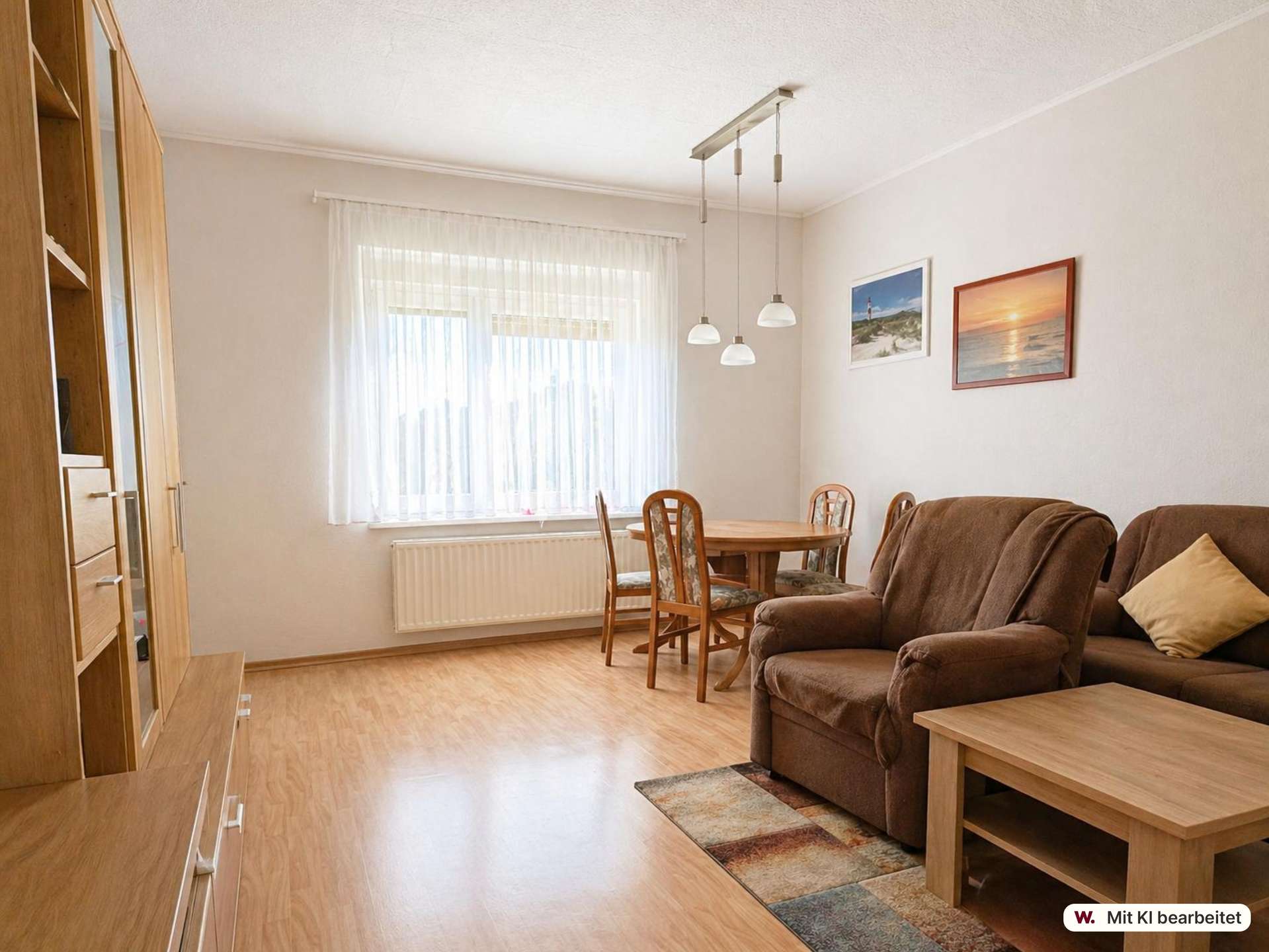 Cozy living-dining room with brown sofa, wooden coffee table, and a round dining table by a window with sheer white curtains; two framed beach photos on the wall.