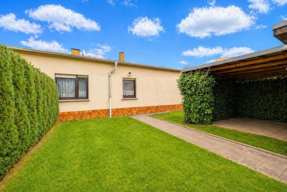 Backyard of a single-story beige house with two windows, a stepped brick base, hedges along the left and a covered patio on the right under a blue sky with clouds