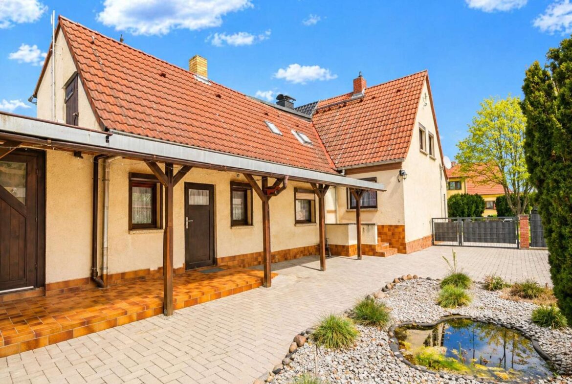 Front view of a beige house with a red tiled roof and wooden porch in a sunny courtyard. A small rock garden with a pond sits in the foreground.