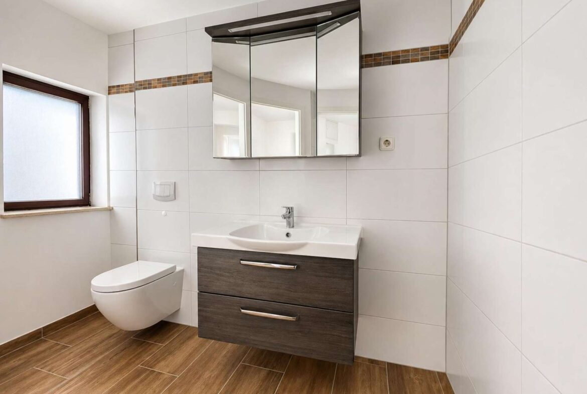 Modern bathroom with a wall-hung toilet, dark wood vanity, and a three-panel mirrored cabinet above a white sink. White tiled walls with brown mosaic trim, and a window to the left.