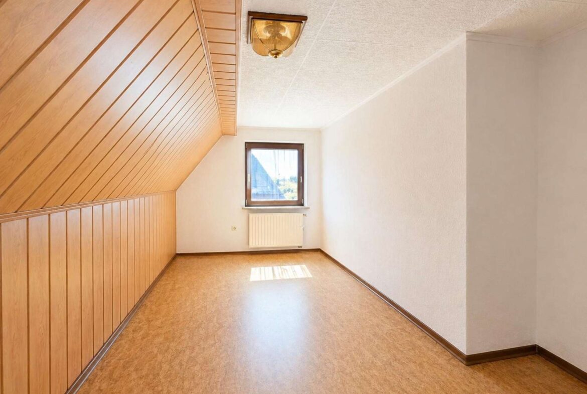 Narrow attic room with wooden paneled slanted wall on left, white walls, small window and radiator beneath it, empty space ahead.