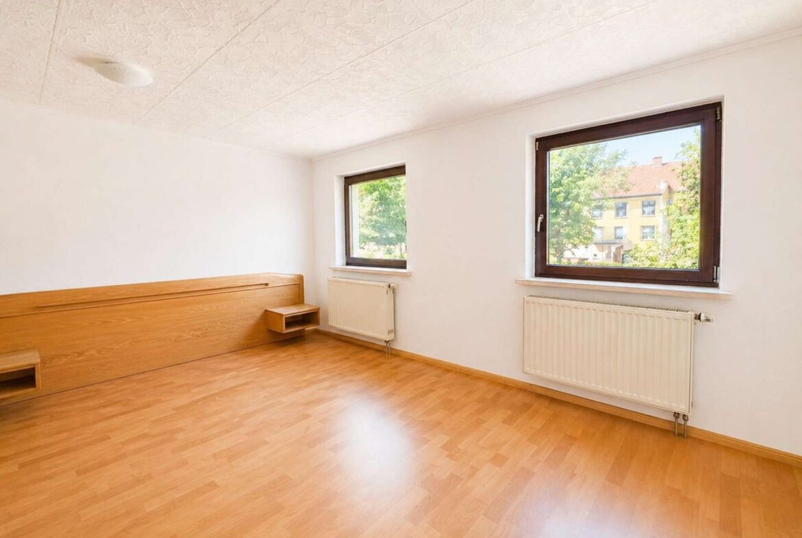 Bright empty bedroom with light wood floor, built-in wooden bed frame, and two large windows..Window view shows trees outside (radiators underneath).