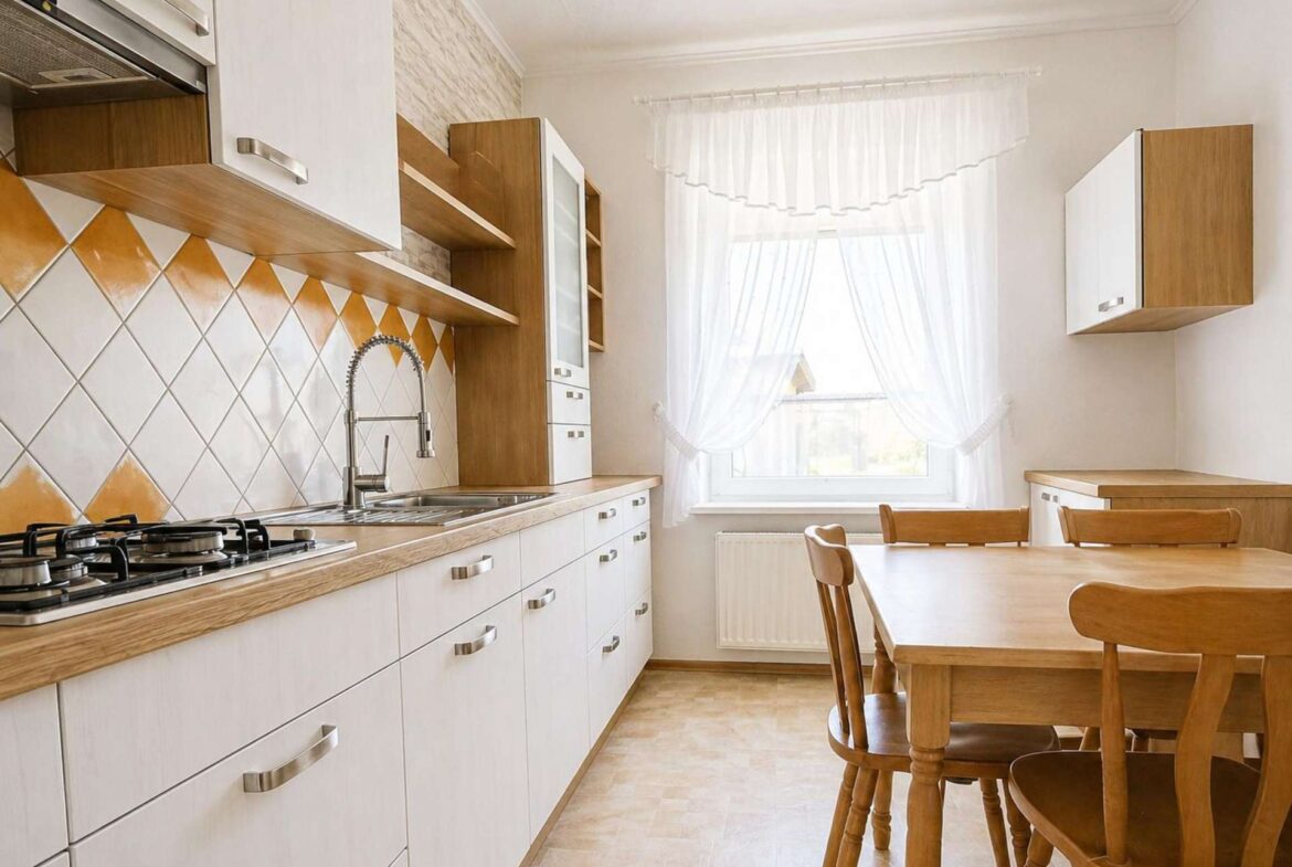 Bright kitchen with light wood cabinets, white countertops, a diamond-pattern orange-and-white tile backsplash, stainless steel sink, and a wooden dining table near a window with sheer curtains.