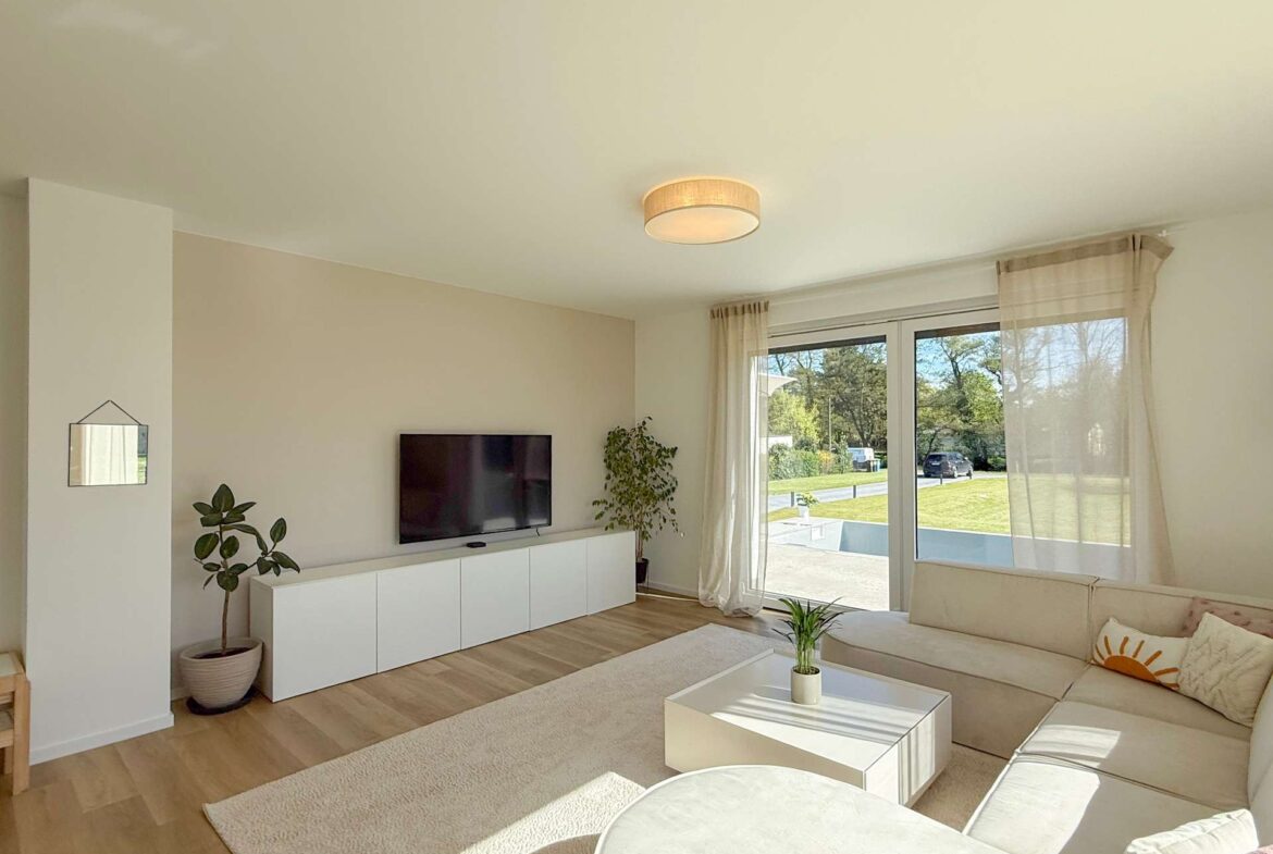 Bright living room with a white TV console and wall-mounted TV, potted plant, and large windows with sheer curtains letting in sunlight