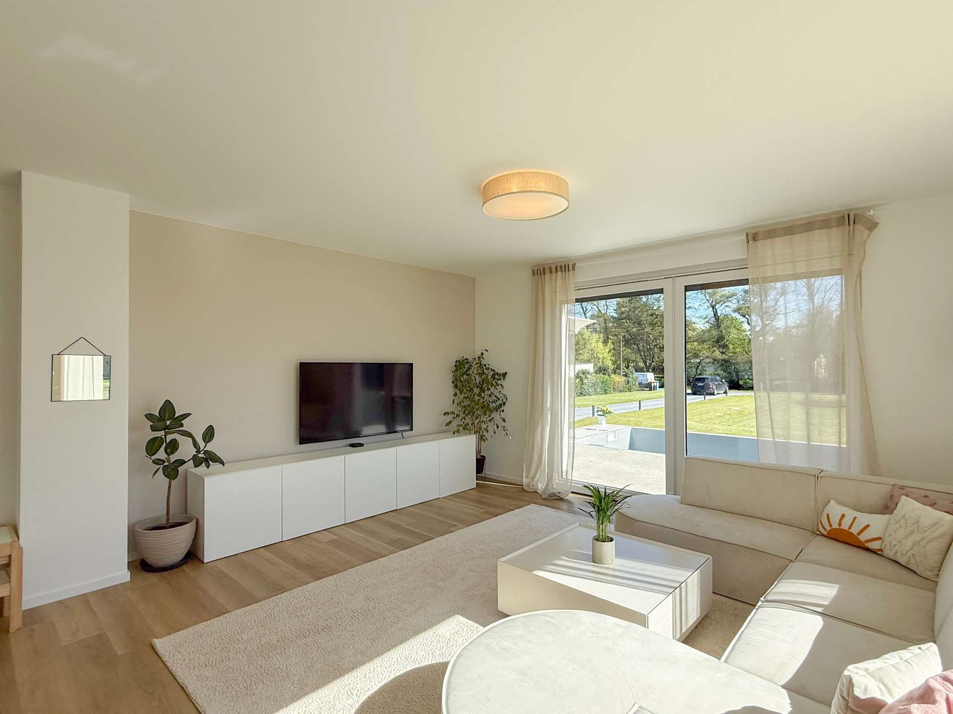 Bright living room with a white TV console and wall-mounted TV, potted plant, and large windows with sheer curtains letting in sunlight