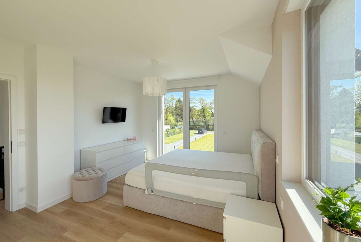 Bright white modern bedroom with a low platform bed, beige headboard, and white dresser; large windows overlook a green lawn outside.