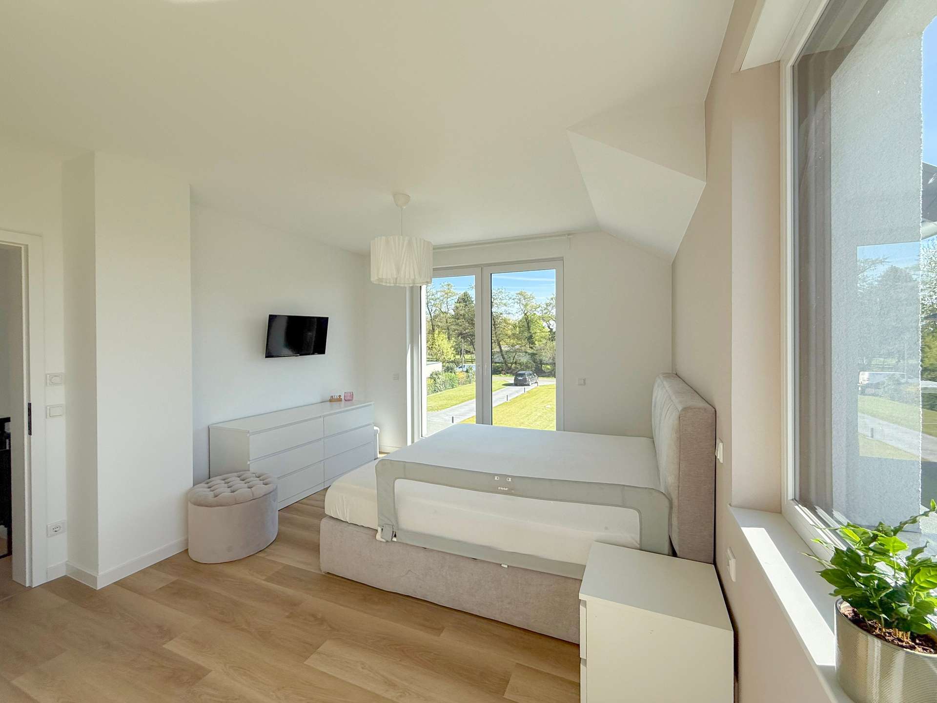Bright white modern bedroom with a low platform bed, beige headboard, and white dresser; large windows overlook a green lawn outside.
