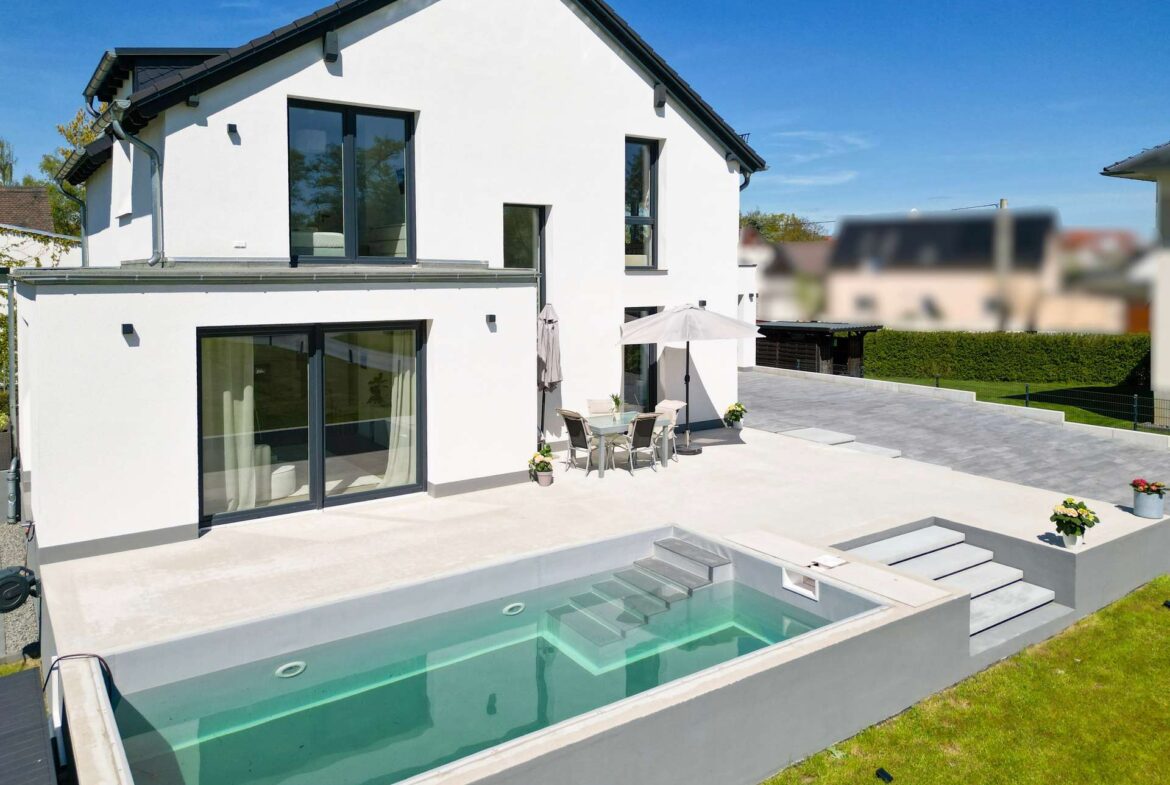 Modern white two-story house with large glass doors and a rectangular pool in the backyard with a patio set nearby.