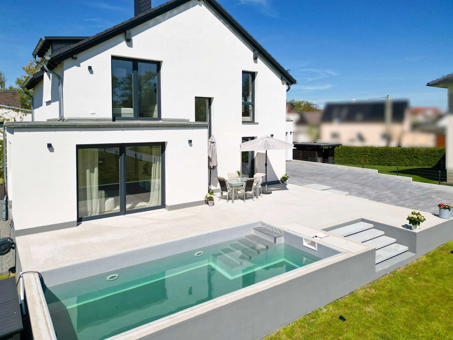 Modern white two-story house with large glass doors and a rectangular pool in the backyard with a patio set nearby.