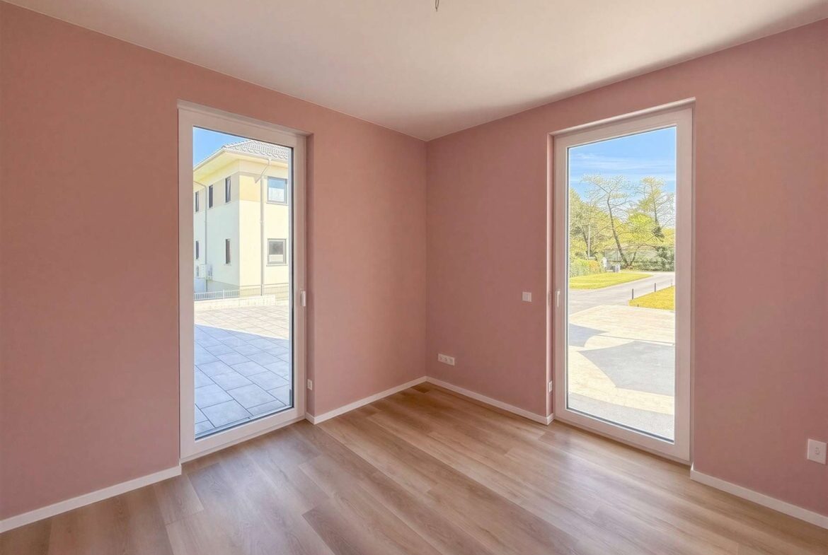 Empty pink-walled room with light wood floor and two large glass doors letting in daylight outside.