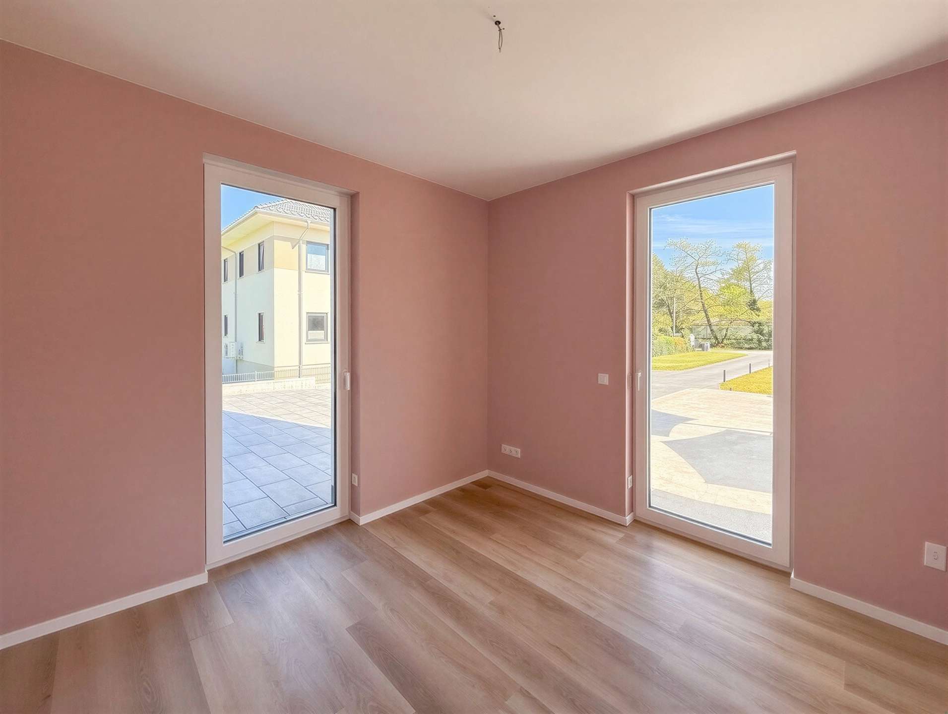 Empty pink-walled room with light wood floor and two large glass doors letting in daylight outside.
