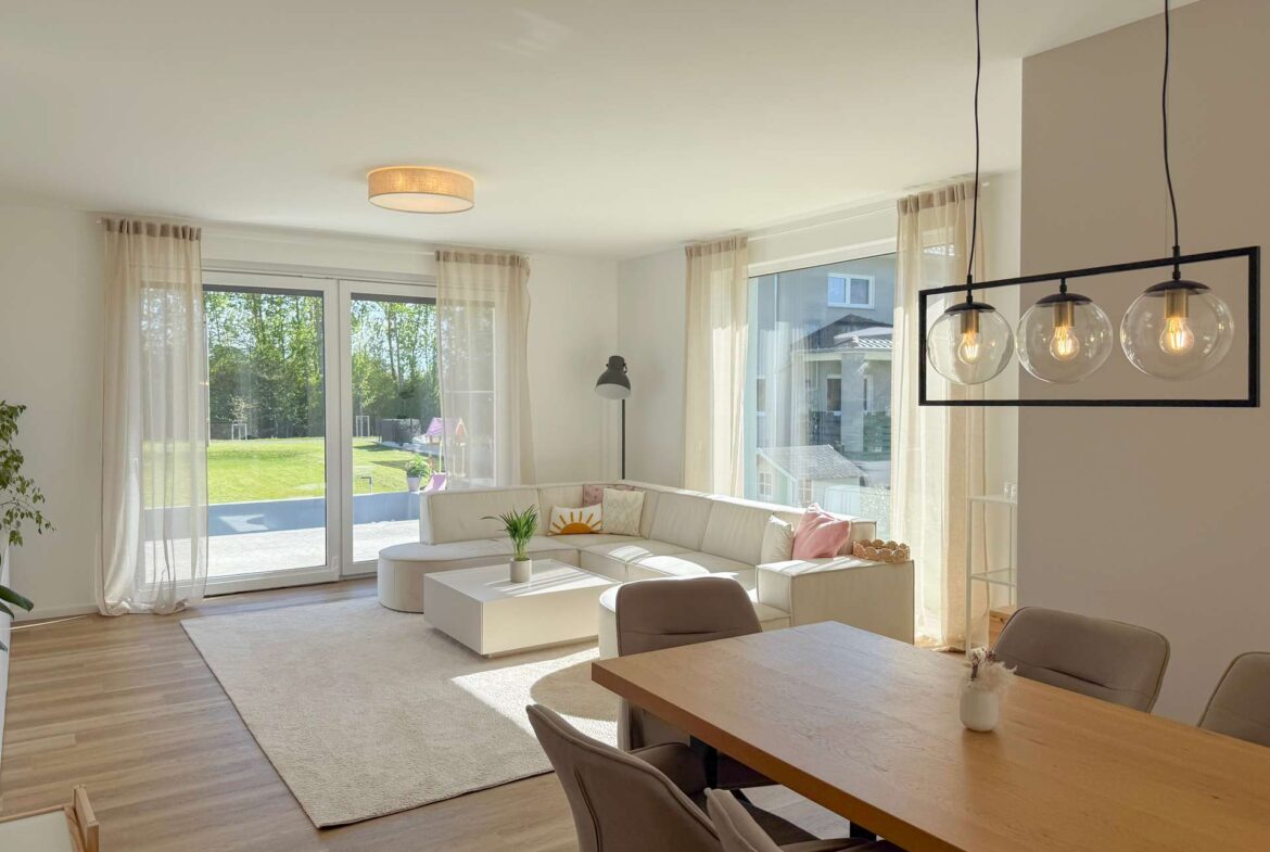 Bright open-plan living room with white sectional sofa, large windows, sheer curtains, and a black-frame hanging light fixture.
