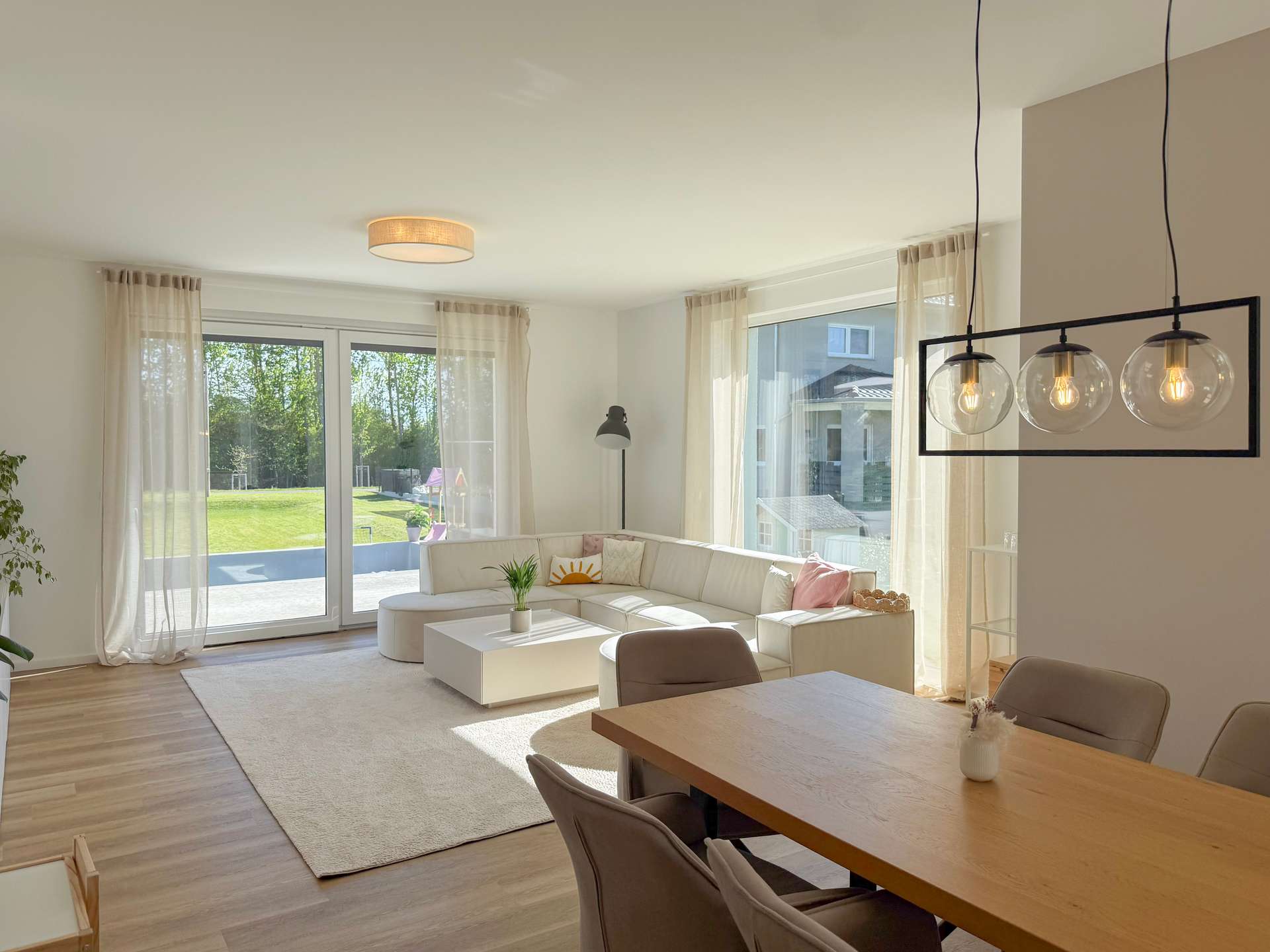 Bright open-plan living room with white sectional sofa, large windows, sheer curtains, and a black-frame hanging light fixture.