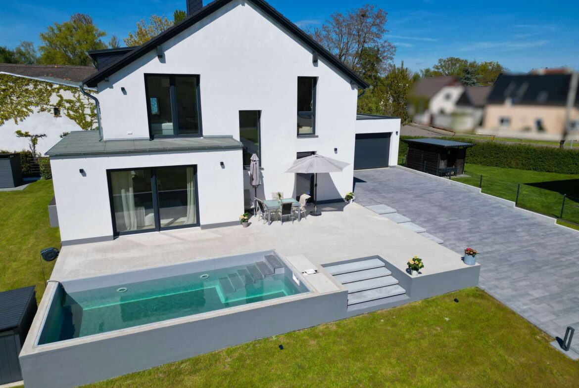 White two-story house with large glass doors, outdoor dining table under an umbrella, and an in-ground pool in the backyard.
