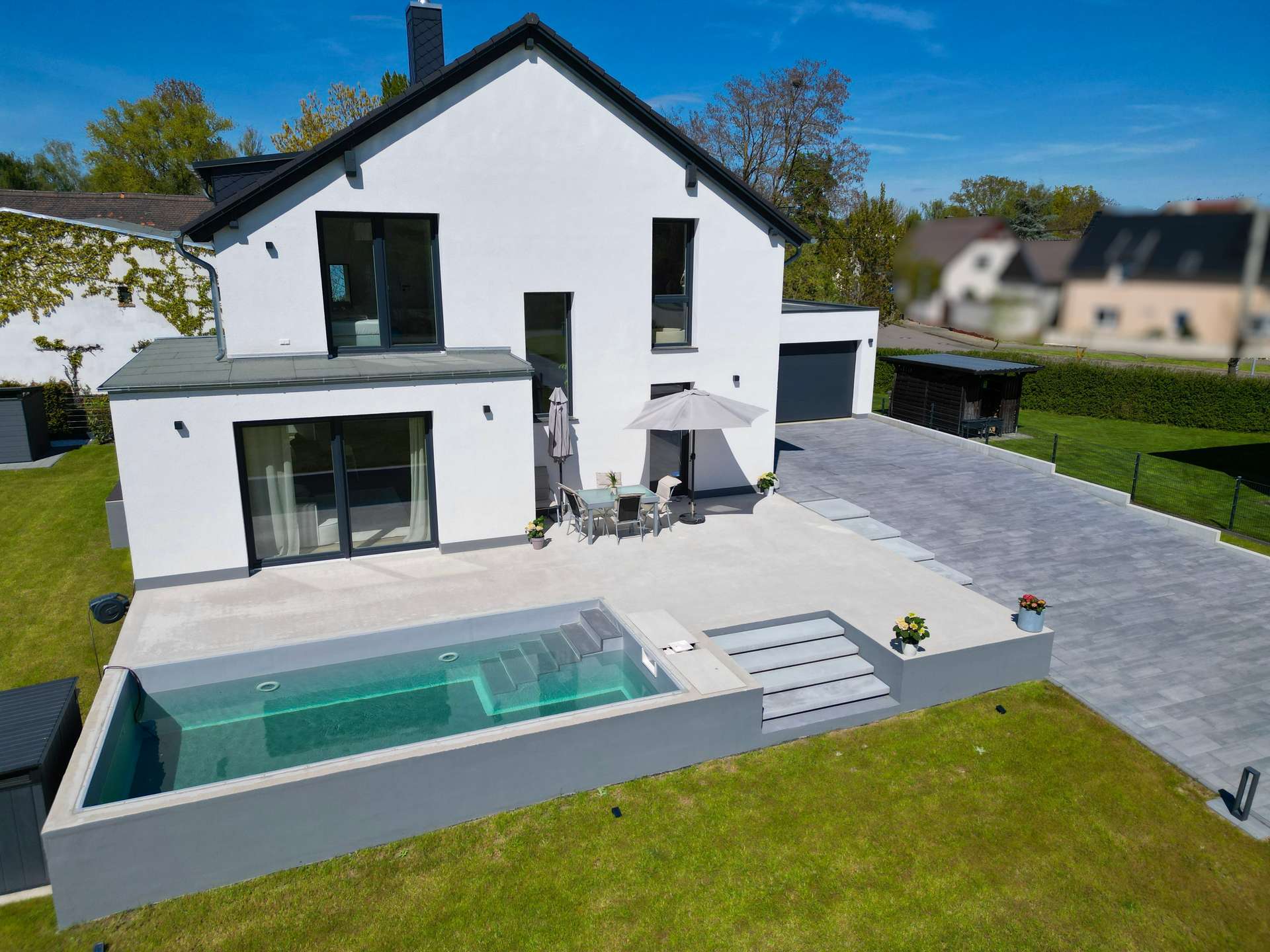 White two-story house with large glass doors, outdoor dining table under an umbrella, and an in-ground pool in the backyard.