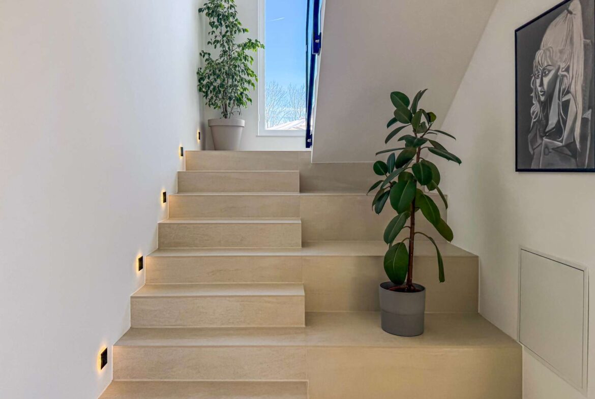 Beige stairway with wall lights along the left, a tall potted plant by the window, and a large leafy plant near a framed artwork on the right wall.