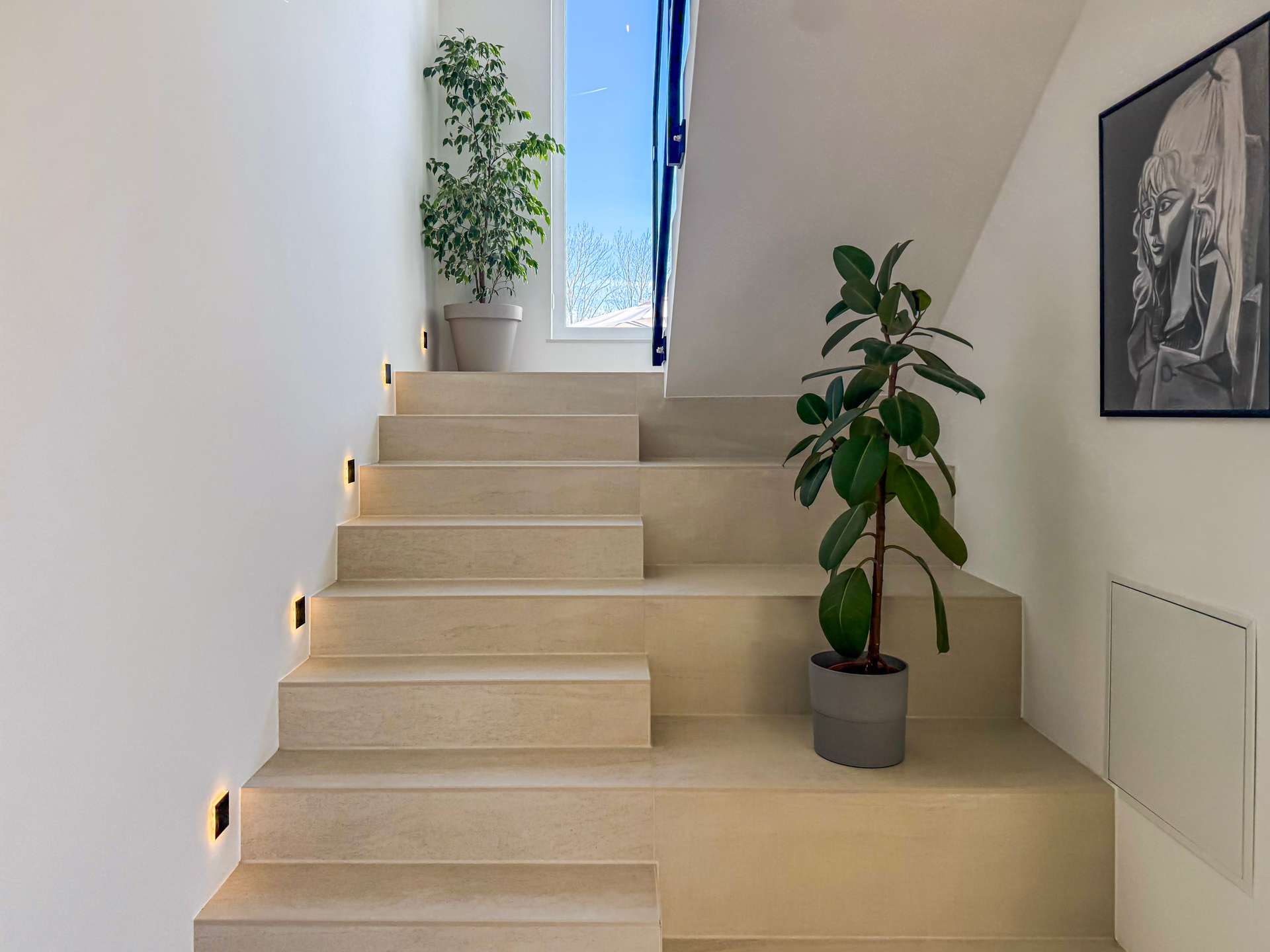 Beige stairway with wall lights along the left, a tall potted plant by the window, and a large leafy plant near a framed artwork on the right wall.