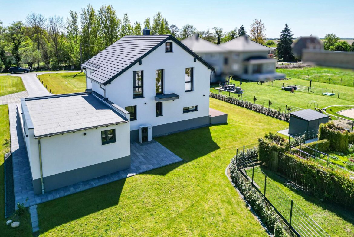 Aerial view of a modern white two‑story house with a dark roof, surrounded by a green lawn and a fenced yard.