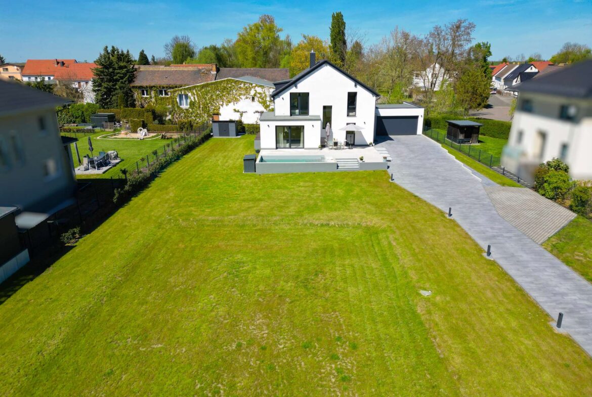 Aerial view of a modern white house with a large green lawn, a gray driveway to a two-car garage, and a rectangular pool area in the yard.