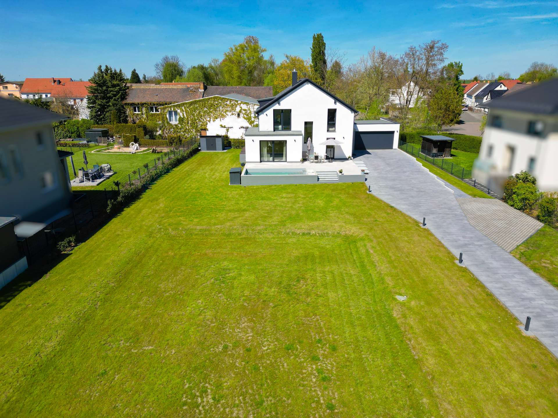 Aerial view of a modern white house with a large green lawn, a gray driveway to a two-car garage, and a rectangular pool area in the yard.