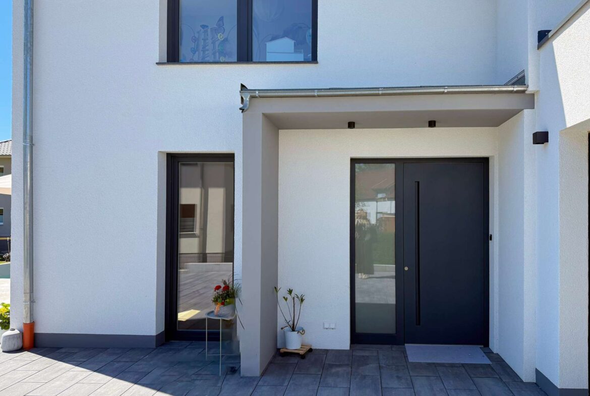 Front entrance of a modern white house with a dark-framed glass door and a large window above, plus potted plants on a gray-tiled porch.