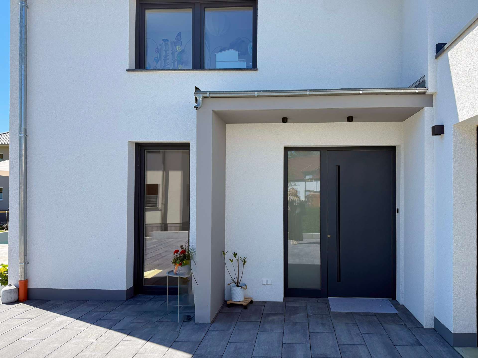 Front entrance of a modern white house with a dark-framed glass door and a large window above, plus potted plants on a gray-tiled porch.