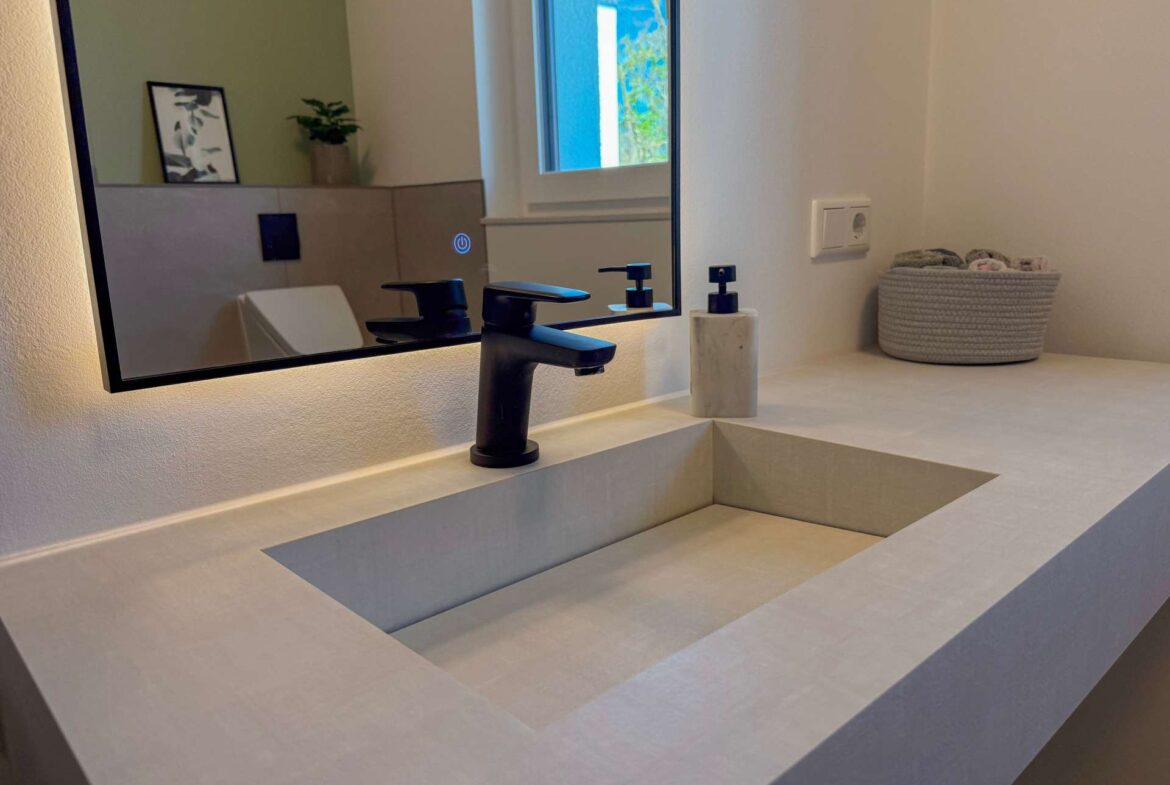 Modern bathroom sink area with a black faucet and marble soap dispenser, mirrored by a large wall mirror reflecting a window and greenery.