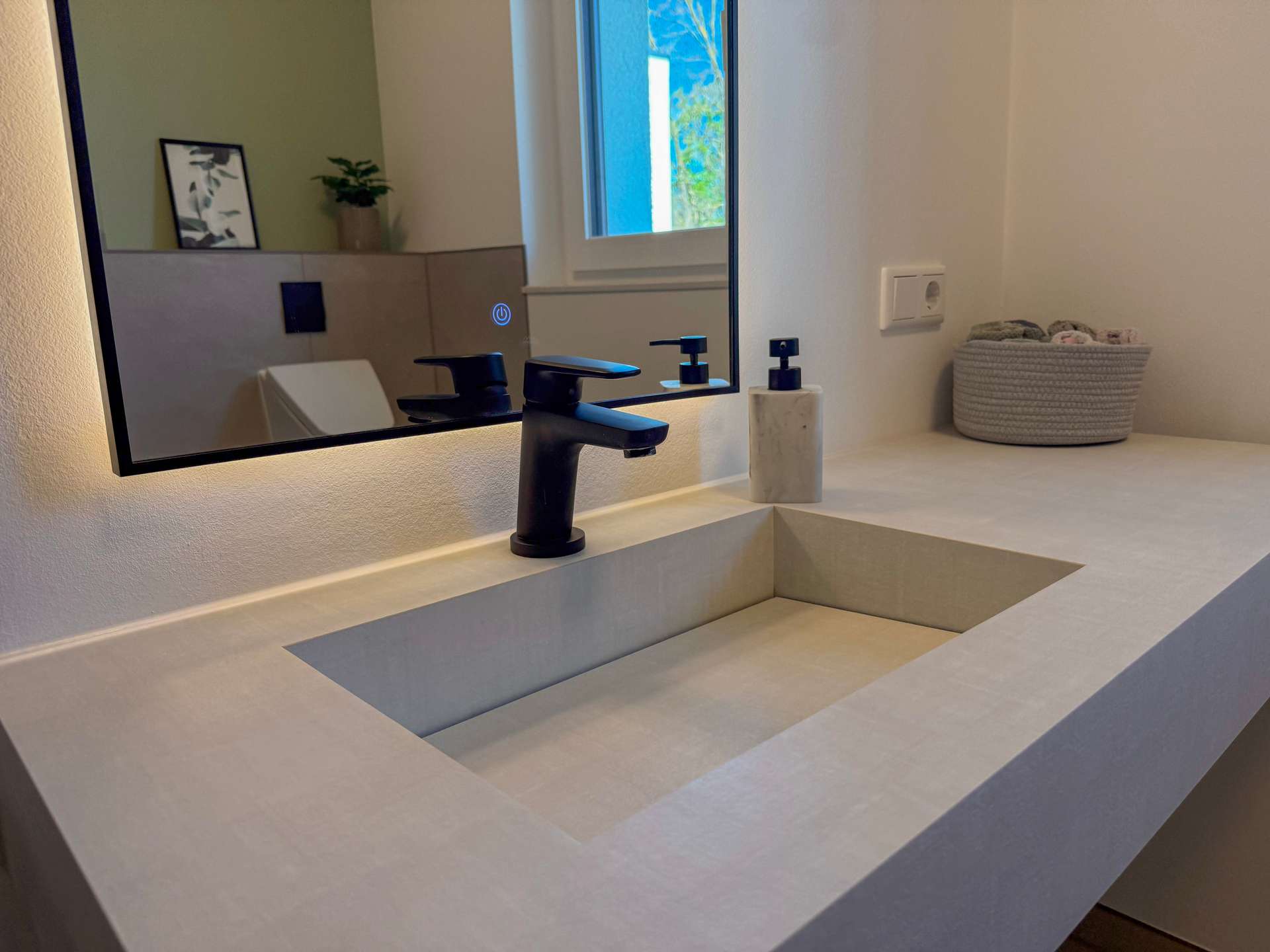 Modern bathroom sink area with a black faucet and marble soap dispenser, mirrored by a large wall mirror reflecting a window and greenery.