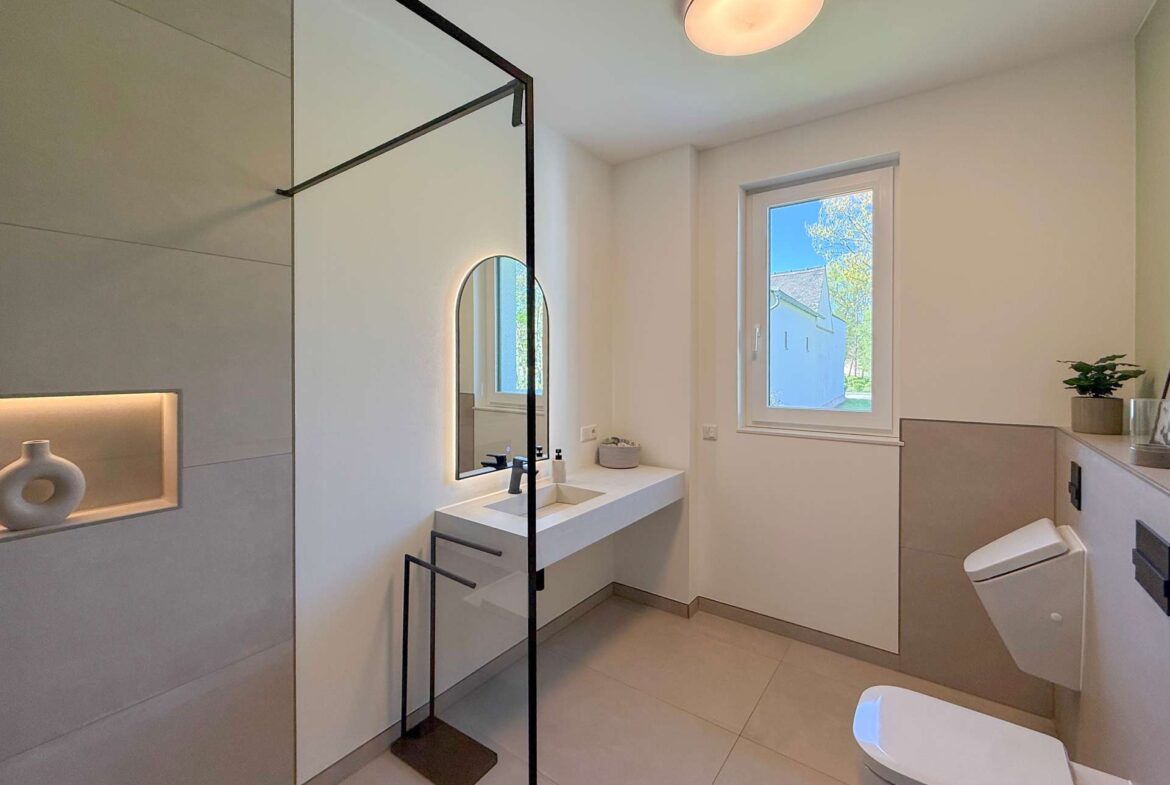 Modern bathroom with glass shower enclosure, floating vanity and oval backlit mirror by a window. A small plant sits on the counter.