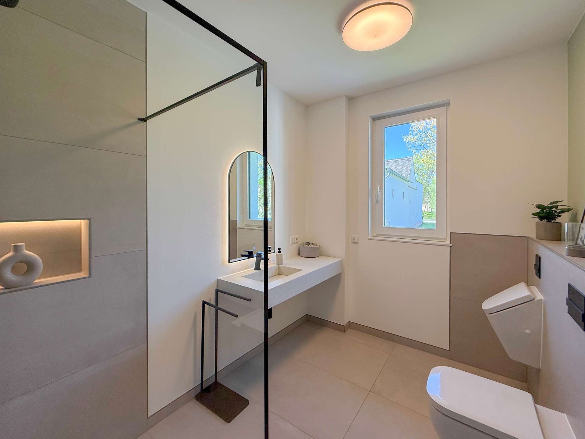 Modern bathroom with glass shower enclosure, floating vanity and oval backlit mirror by a window. A small plant sits on the counter.