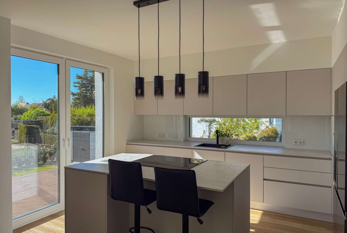 Modern kitchen with an island, four pendant lights, and a large window overlooking a garden.