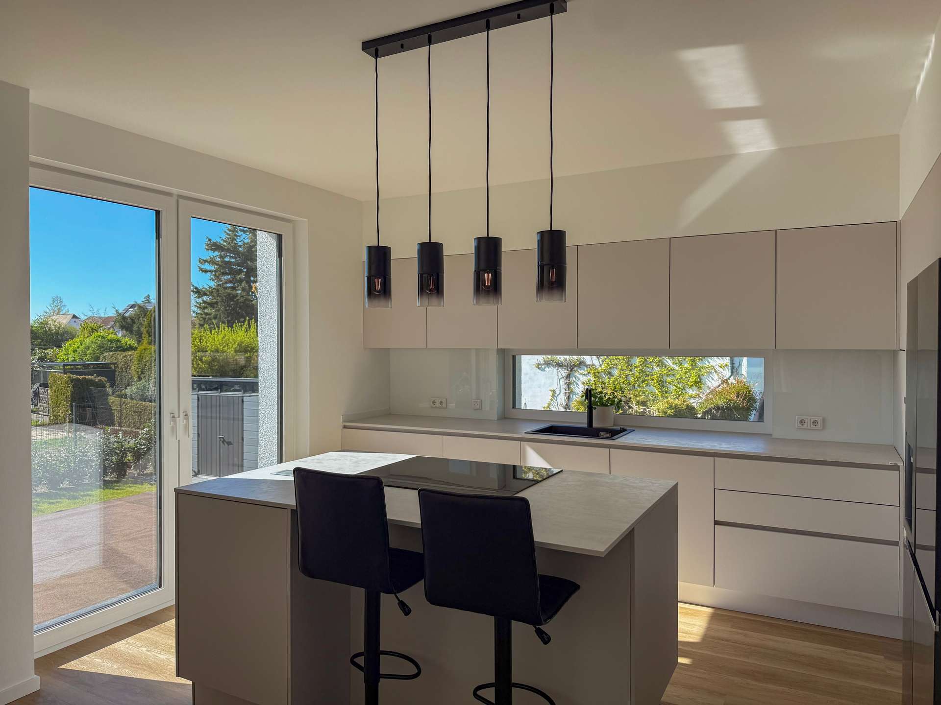 Modern kitchen with an island, four pendant lights, and a large window overlooking a garden.