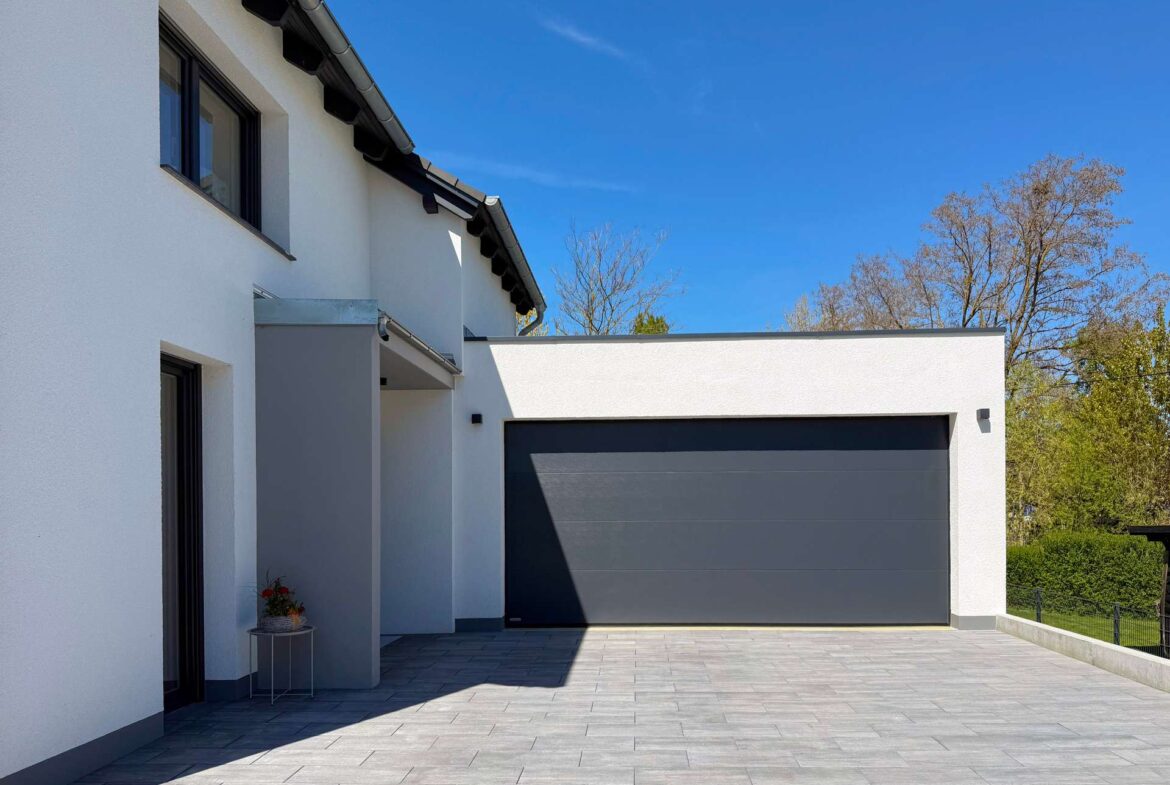 Modern house exterior with white walls and a large dark grey garage door; tiled driveway and clear blue sky.