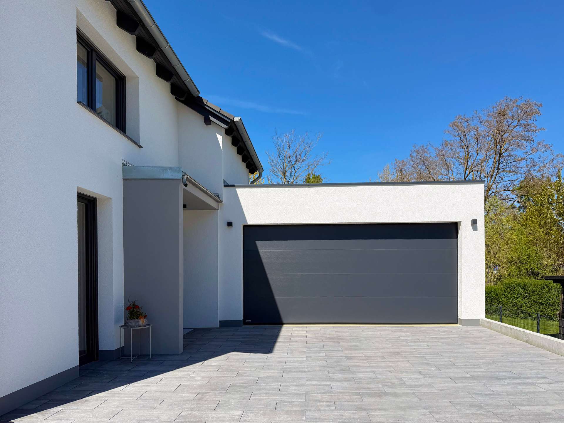 Modern house exterior with white walls and a large dark grey garage door; tiled driveway and clear blue sky.