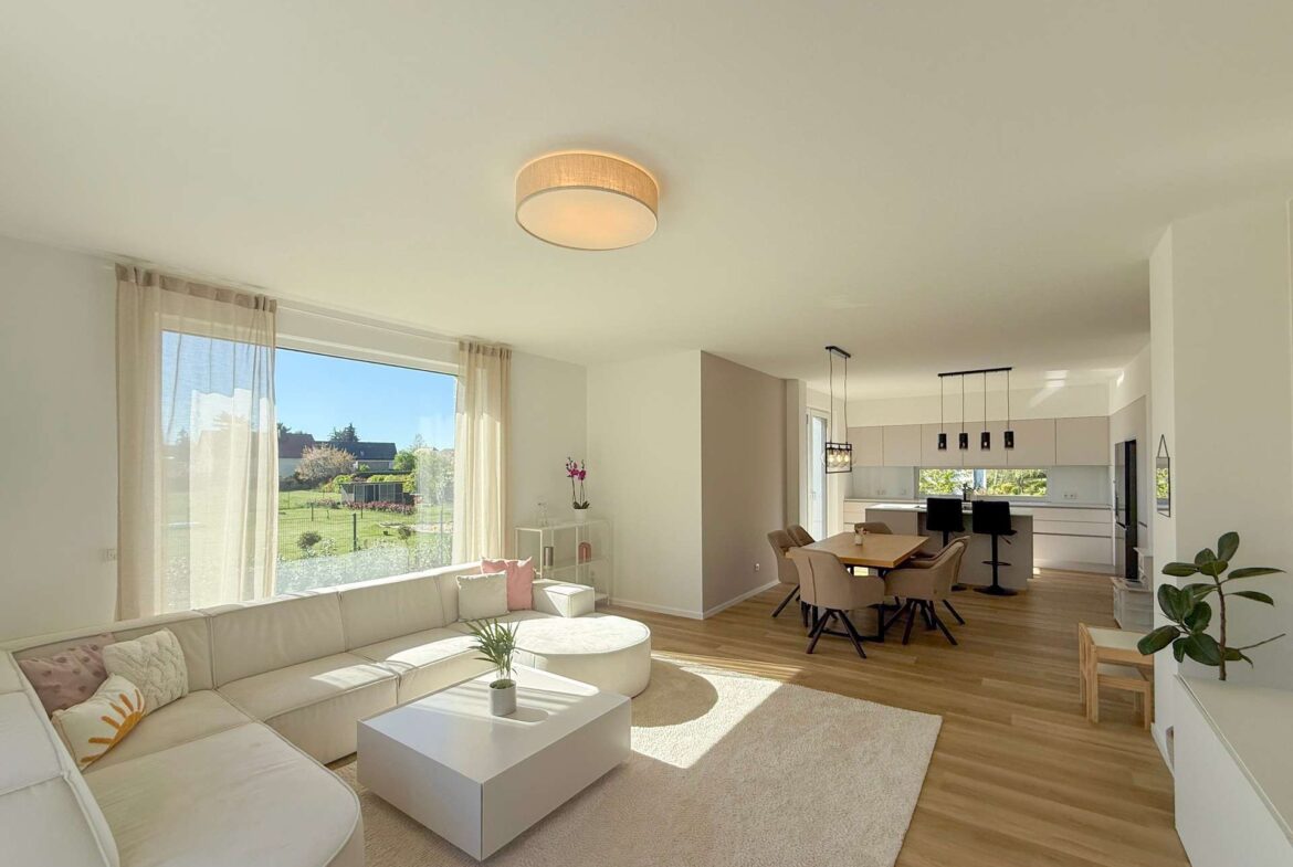 Bright open-plan living room and kitchen with a white sectional sofa, beige curtains, and a large window overlooking a garden.