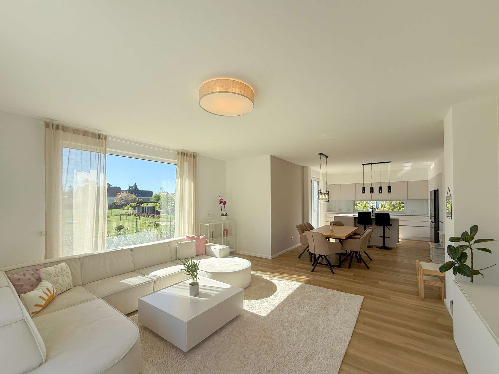 Bright open-plan living room and kitchen with a white sectional sofa, beige curtains, and a large window overlooking a garden.