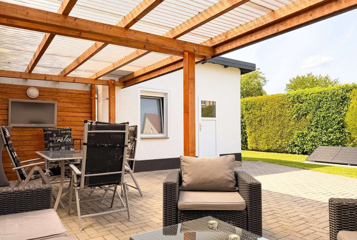 Covered patio with wooden beams and translucent roof, outdoor wicker furniture and glass coffee tables in a sunny backyard setup.