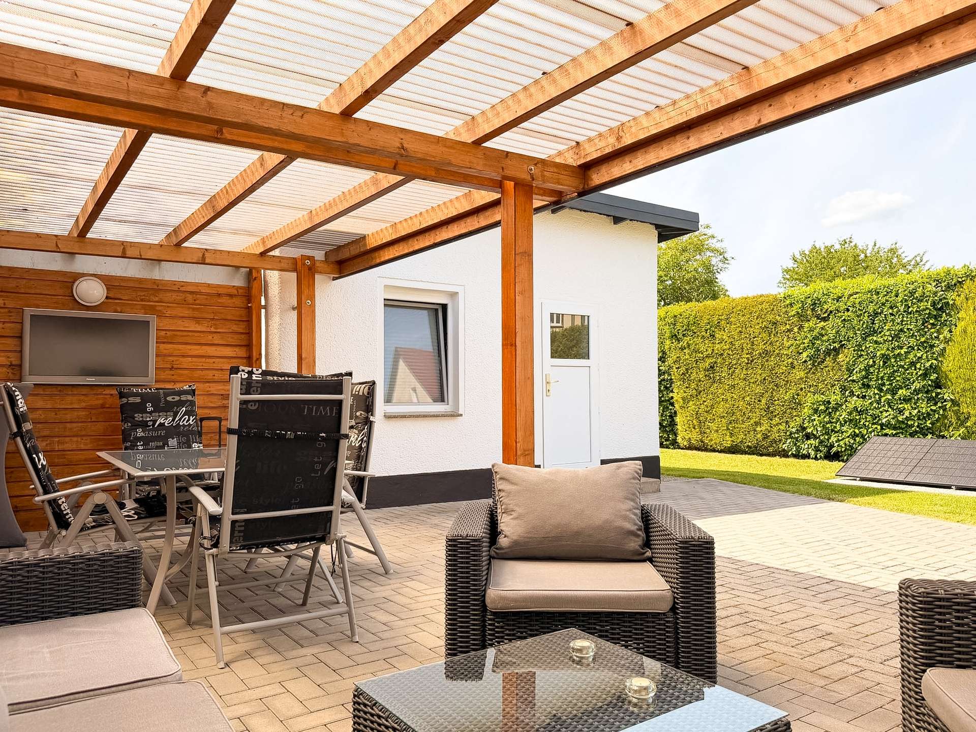 Covered patio with wooden beams and translucent roof, outdoor wicker furniture and glass coffee tables in a sunny backyard setup.