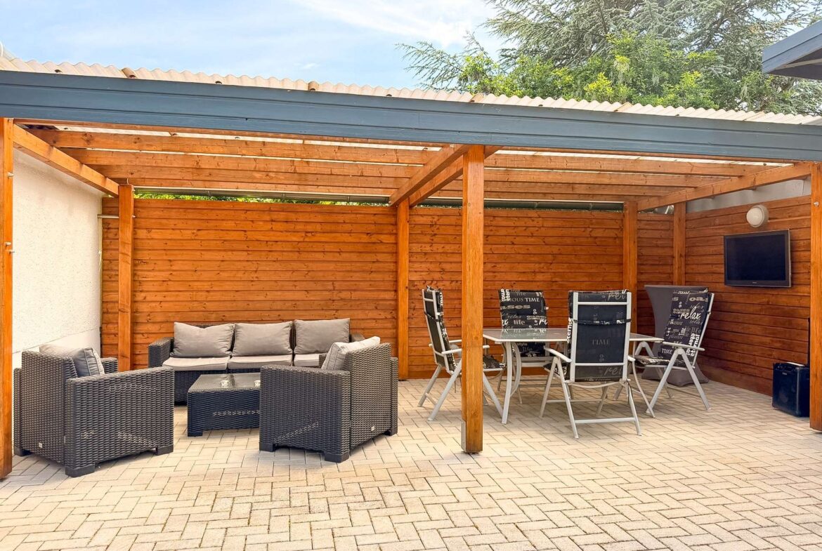 Outdoor patio with a cushioned wicker sofa set under a wooden pergola and a glass coffee table on a brick-paved floor.