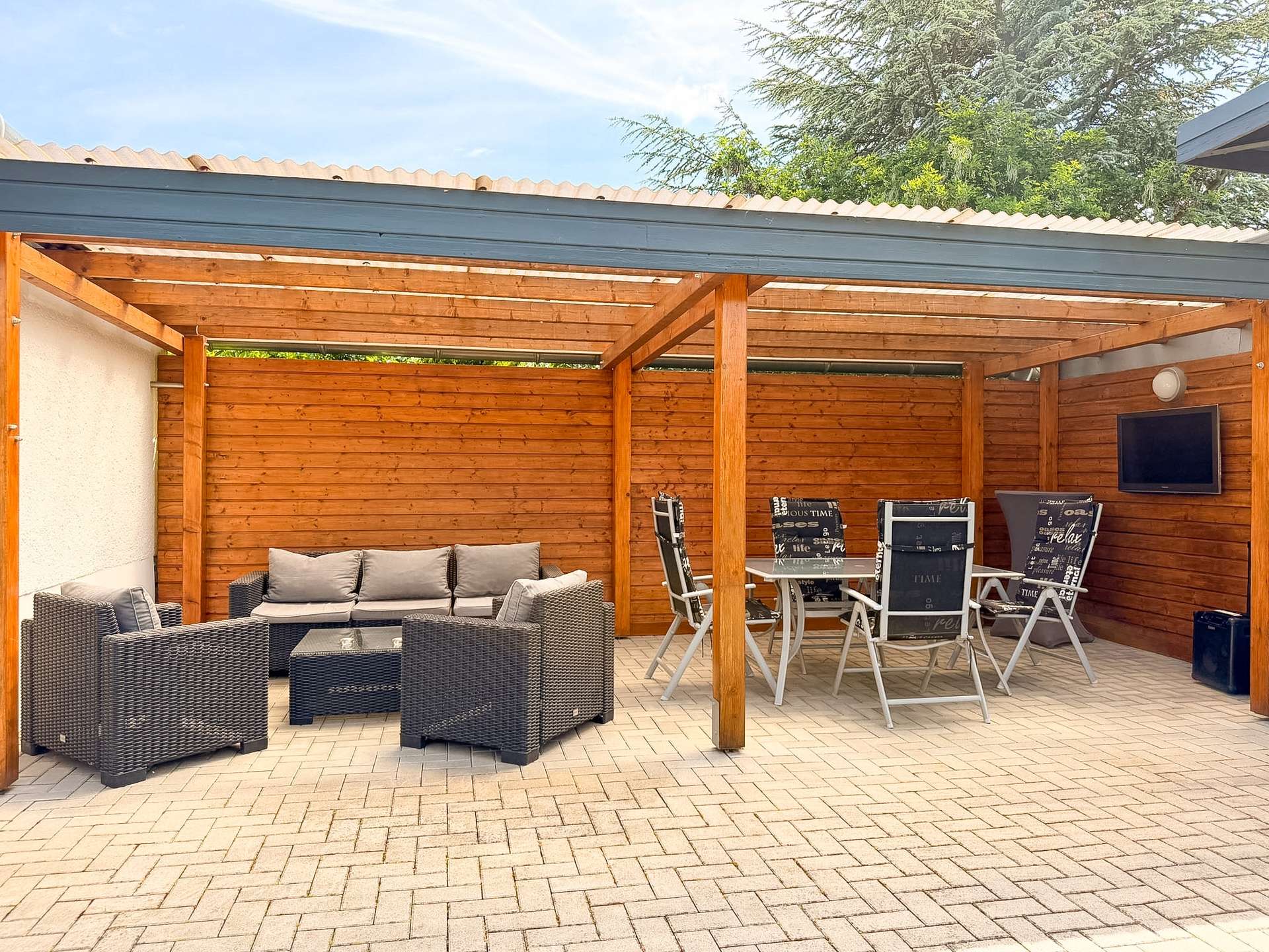 Outdoor patio with a cushioned wicker sofa set under a wooden pergola and a glass coffee table on a brick-paved floor.