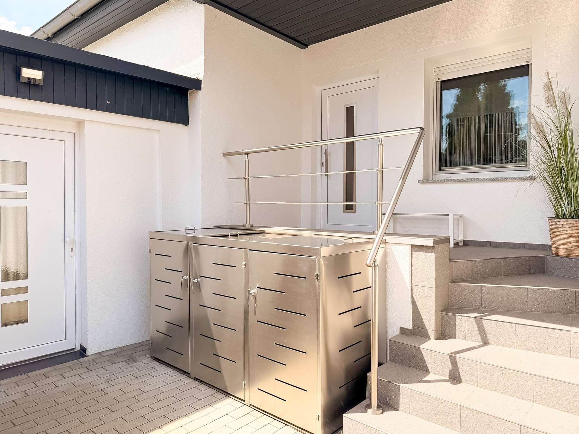 Outdoor stainless steel barbecue grill cabinet beside entry stairs on a modern white house porch.