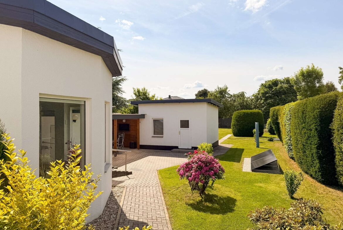 Modern white houses with a paved path through a tidy garden under a sunny, blue sky surrounded by trimmed hedges and colorful shrubs.