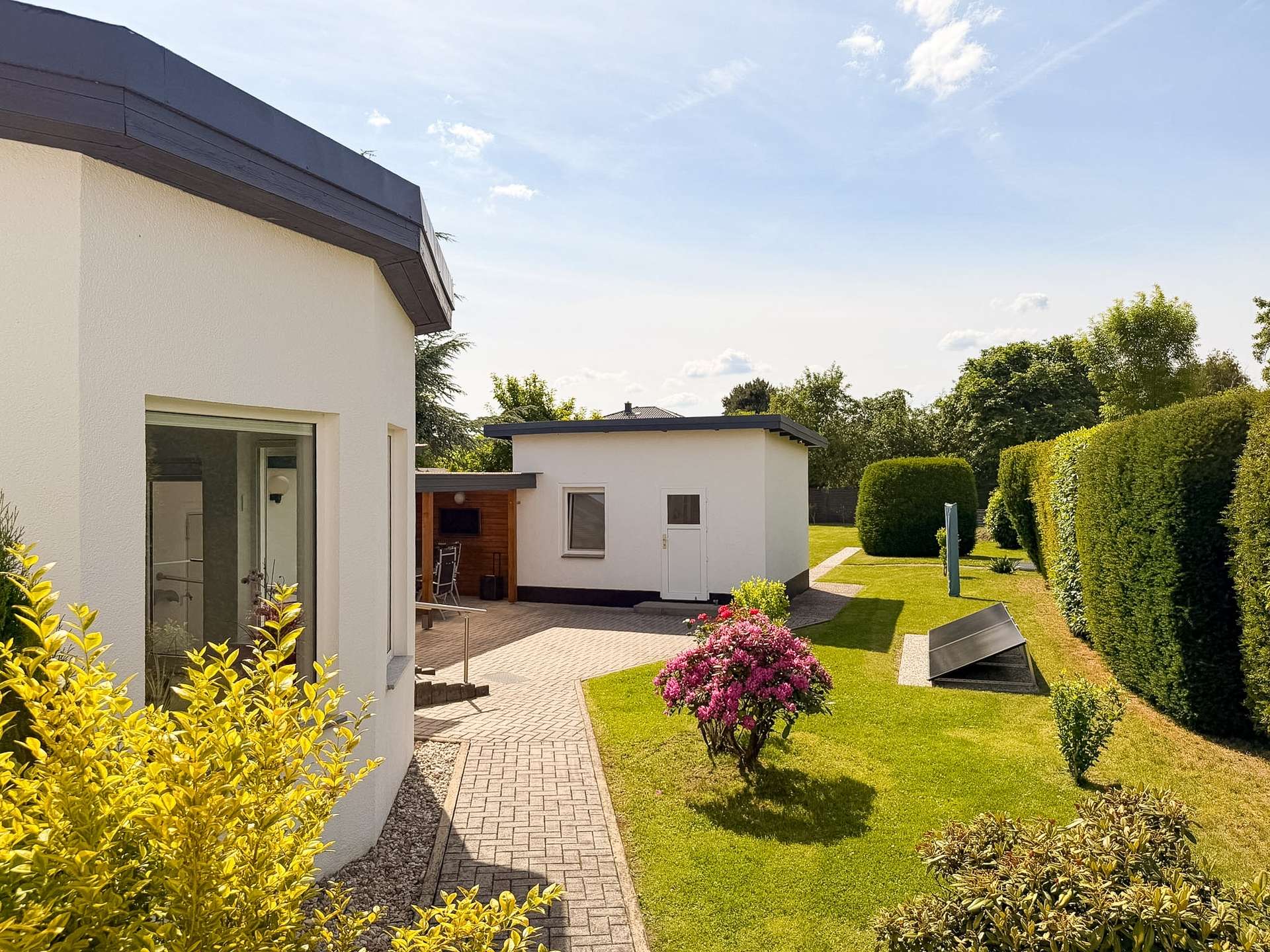 Modern white houses with a paved path through a tidy garden under a sunny, blue sky surrounded by trimmed hedges and colorful shrubs.
