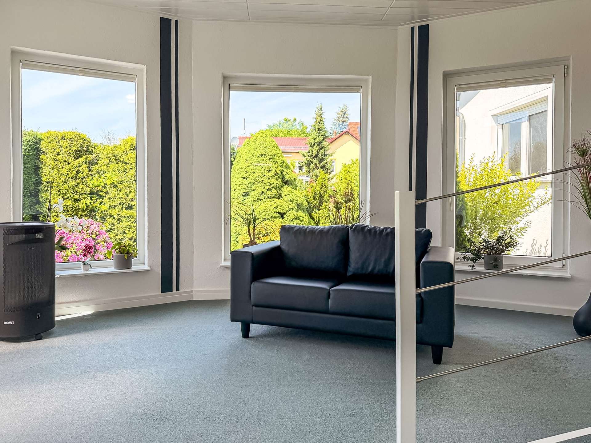 Bright living room with three large windows, a dark blue sofa, and potted plants on the window sills; a speaker visible on the left side of the room.