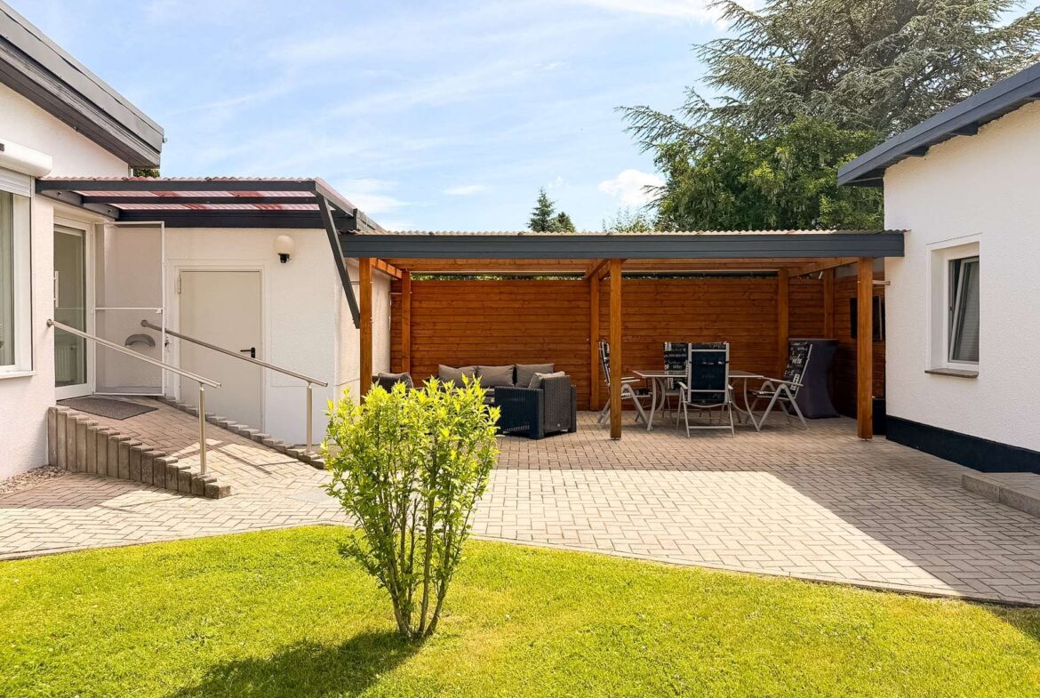 Outdoor patio area with wooden pergola, seating, and a glass table set against white walls of a house. A green lawn and bright sky surround the space.