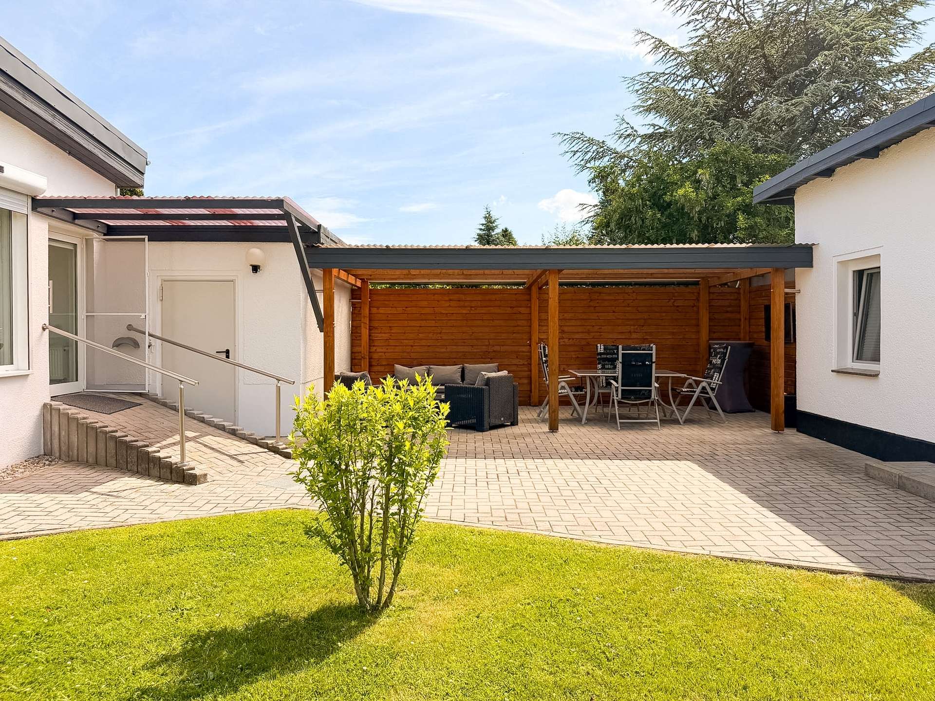 Outdoor patio area with wooden pergola, seating, and a glass table set against white walls of a house. A green lawn and bright sky surround the space.