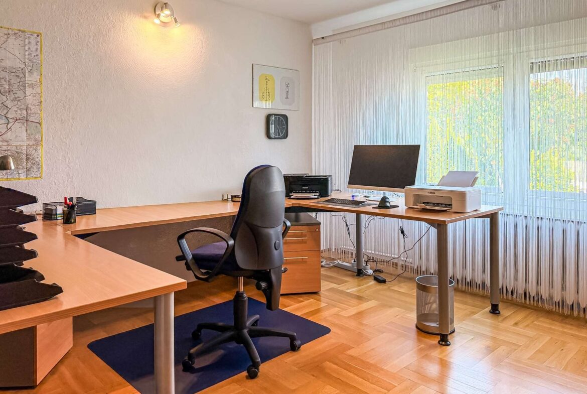 Bright corner office with L-shaped desks, black swivel chair, computer monitor, printer, and large window with vertical blinds, daylight flooding the room.