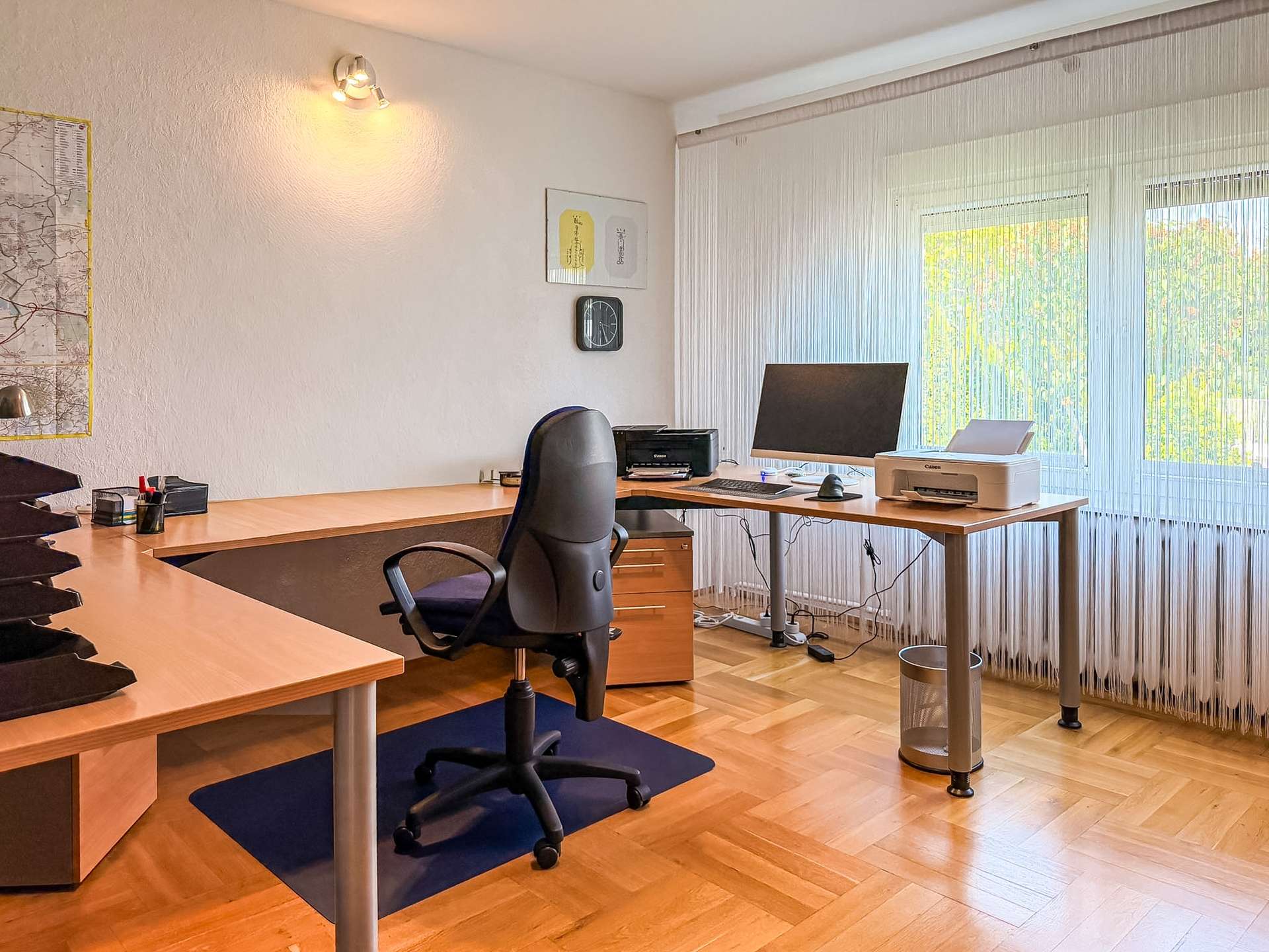Bright corner office with L-shaped desks, black swivel chair, computer monitor, printer, and large window with vertical blinds, daylight flooding the room.