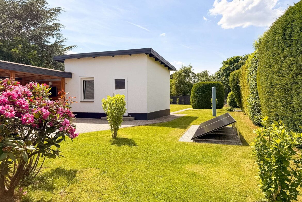 White single-story house in a sunny yard with a pink flowering shrub in the foreground and solar panels on the ground nearby.
