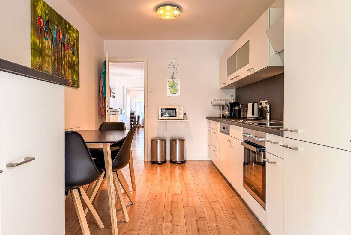 Bright modern galley kitchen with white cabinets, stainless steel appliances, a small dining table and black chairs at the left.